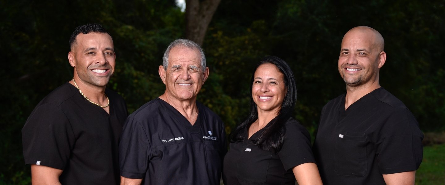 A group of four dental professionals wearing black scrubs, standing outdoors with trees in the background. They are smiling, and one of them has Dr. Jeff Collins embroidered on his uniform. | Collins & Sampson Family & Cosmetic Dentistry