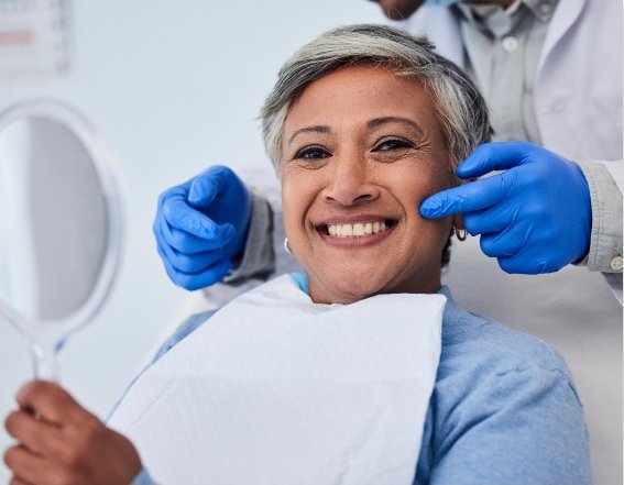A mature woman with short gray hair sits in a dental chair, holding a mirror and smiling. A dentist wearing blue gloves points towards her face, indicating a successful dental treatment. | Collins & Sampson Family & Cosmetic Dentistry