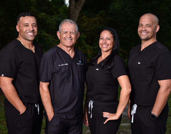A group of four dental professionals wearing black scrubs, standing outdoors with trees in the background. They are smiling, and one of them has 